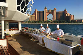Group of tourists dining on a luxury yacht deck with Atlantis The Palm hotel visible across the water in Dubai